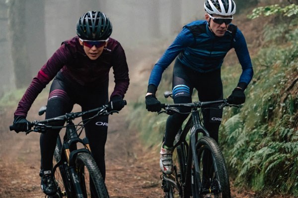 Two mountain bikers wearing bib-shorts with leg warmers and long-sleeve jerseys with base layers