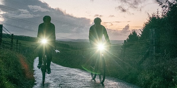 two cyclists riding with lights at dusk on a country road