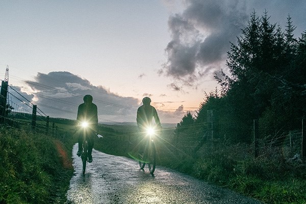 Two cyclists on country lanes with lights at dusk