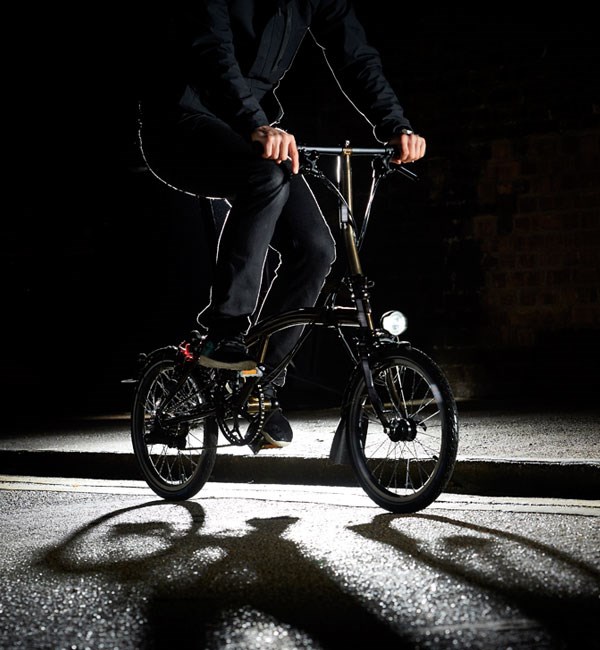 A cyclist rides a Black Edition Brompton with Rocket Red frame along dark streets